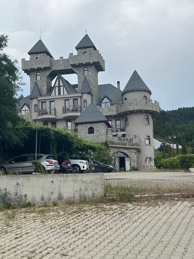 The image shows a castle-like building with multiple towers, crenellations, and arched windows. The building is constructed of stone and stucco and appears to be a hotel or resort. In front of the building, there is a parking area with several cars. A wall separates the parking area from a paved walkway with a patterned surface. Greenery, including trees and vines, surrounds the building. The sky is overcast. The building is identified as the Royal Castle hotel in Ognyanovo, Bulgaria.