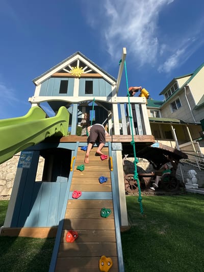 In the city of Danilovka (Altai), Russia, a sun-drenched outdoor scene unfolds featuring a multi-level wooden playground. The playground, painted in shades of blue and yellow with a green slide, is the central focus. A young boy, wearing brown shorts, is actively climbing the playground's wooden ladder with colorful climbing holds. Another child, partially obscured, stands on the playground's upper level. To the right, a yellow telescope is held by someone wearing an orange hat. In the background, a light-colored building with a green roof is visible. Below the playground, a woman in a green dress sits on a bench under a rustic awning, surrounded by decorative wagon wheels. A stone wall and a manicured lawn complete the environment, all under a bright blue sky with scattered clouds.
