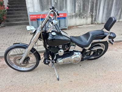 The image shows a black and chrome Harley-Davidson motorcycle parked on a paved surface. A building with a set of stairs and a gray concrete wall are behind the motorcycle. There is a red and white “CEPSA” branded enclosure behind the motorcycle. The environment appears to be outdoors during daylight hours. The precise location within Soure, Portugal, is not discernable from the image.