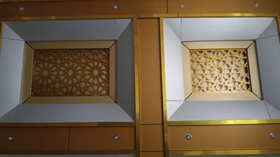 The image shows a close-up view of decorative panels on a wall, likely an interior space. The panels feature a repeating geometric pattern in gold, reminiscent of Islamic art, set within recessed frames. The frames themselves are outlined in a metallic gold trim, and the surrounding wall elements are a mix of neutral colors, including a pale grey and a muted orange-brown. The overall impression is one of modern design with traditional influences. The scene is static and does not depict any people or actions. There is no visible indication of its location in Julikeude Dua, Indonesia.