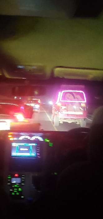 The image depicts a night scene observed from the interior of a vehicle on a road in Naivasha East ward, Kenya. The foreground shows the illuminated dashboard, including a digital infotainment display and climate control buttons, within the observing vehicle. Ahead, on a multi-lane paved road, traffic is visible. Prominently in the immediate foreground traffic is a light-colored utility vehicle, potentially pink or red-orange, with elevated ground clearance and visible mud on its lower rear. This vehicle is custom-lit with bright magenta/pink neon-like outlining around its rear window. A large, illuminated decal featuring a stylized depiction of a person, likely a woman, is displayed within the rear window of this vehicle. Further along the road, additional vehicles are discernible primarily by their red taillights and distant white headlights. The environment is characterized by artificial illumination against a dark backdrop.