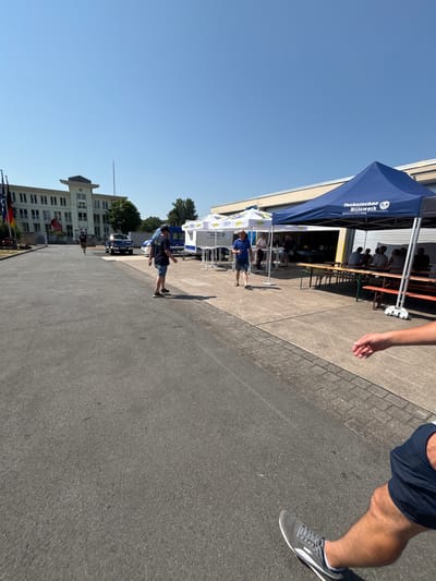 The image captures an outdoor gathering in Obernburg am Main, Germany, on a clear, sunny day. In the background, a large, modern building with multiple windows stands under a bright blue sky. Adjacent to the building, to the right, is a blue tent with the inscription "Technisches Hilfswerk" and "Ortsverband Obernburg". Several white tents are set up in front of the building, some displaying logos. Long tables with benches are arranged under the tents, suggesting a communal eating or gathering area.
Several people are visible in the scene. Two men are walking on the paved area in the foreground, one wearing a blue shirt and shorts, the other in a dark t-shirt and shorts. More people are seated at the tables under the tents, engaged in conversation. The ground is a mix of asphalt and paved cobblestone-like patterns. Flags are visible near the entrance of the building on the left. A dark blue pick-up truck and a white van are parked near the tents. The overall atmosphere appears to be a casual outdoor event. The image is taken from a low-angle perspective, with the lower legs and feet of a person visible in the right foreground, suggesting the photographer's presence.