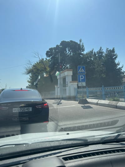 A dark sedan is driving down a street in Baytkurgan, Uzbekistan. The car has a license plate that reads "01 0705 MB UZ." In the background, there is a traffic light, a pedestrian crossing sign, a parking sign, and a sign for disabled parking. There are also trees and a blue fence. The sky is clear and blue.