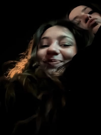A photograph in low light features two faces. The primary face, angled, has light skin, brown hair, and a slight smile. The features are somewhat blurred. A second face, smaller and darker, is located in the upper left corner. The background is entirely black. Light glints off the ends of the primary figure’s hair. The overall impression is one of motion or a distorted perspective.