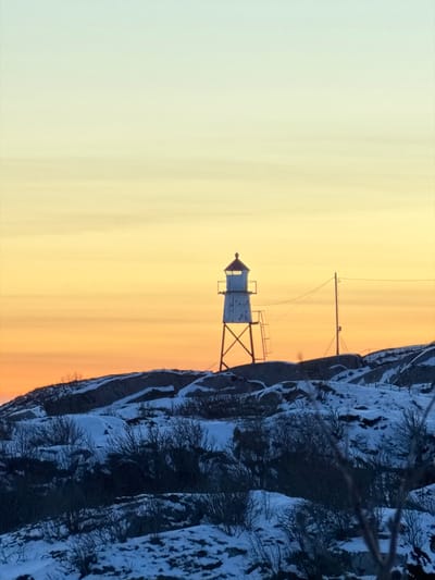 A lighthouse sits atop a snowy cliff against a gradient sky. The lighthouse is white with a dark roof, supported by a metal frame. A taller antenna is visible nearby. Snow covers the rocky cliff face, interspersed with dark trees and branches. The sky transitions from a pale yellow near the horizon to a light blue above. The photo is oriented vertically.