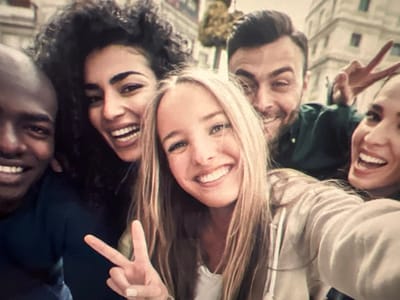 Five smiling individuals pose closely together, seemingly taking a selfie. A Black man on the left and a woman with curly hair next to him smile broadly. A blonde woman in the center flashes a peace sign. Behind her and slightly to the right, a man makes a peace sign while grinning. Finally, a woman is seen on the far right, also with a wide smile. The background is blurry, suggesting an urban environment. The overall atmosphere is cheerful and friendly.
