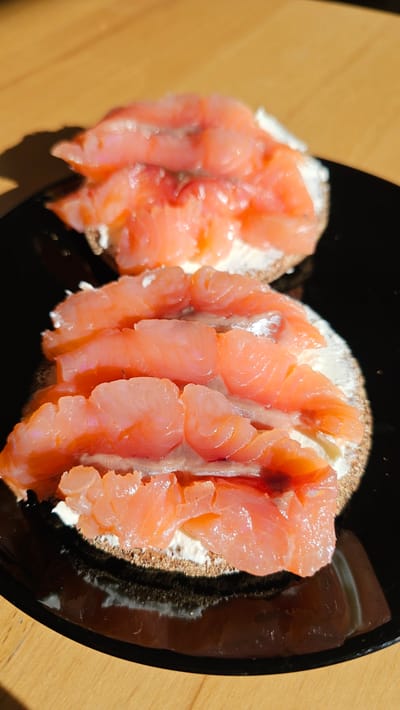 A top-down close-up shot shows two halves of a bagel topped with cream cheese and smoked salmon. The bagel halves are on a black plate, which is on a wooden table. The salmon slices are a vibrant pink-orange and are arranged in rows on the cream cheese. The cream cheese is white and appears thick. The bagel has a brown crust. The lighting suggests it is daytime, with a shadow cast on the table to the left of the plate, indicating the sun is coming from the right. The scene is simple and focused on the food, creating an appetizing visual. There is no visible text in the image. The setting appears to be indoors, possibly a kitchen or dining area, given the wooden table and indoor lighting. The overall impression is of a fresh and ready-to-eat meal.