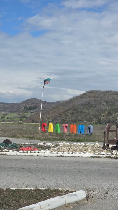 The image depicts a roadside scene in Slatino, Bulgaria. The name "SLATINO" is spelled out in large, colorful, individual letters positioned on the side of a road, bordered by white-painted rocks. To the left, a Bulgarian flag flies on a pole. In the background, a hillside covered with sparse vegetation can be seen under a cloudy sky. A gray asphalt road is located to the right. A white concrete curb is located on the right side of the road.