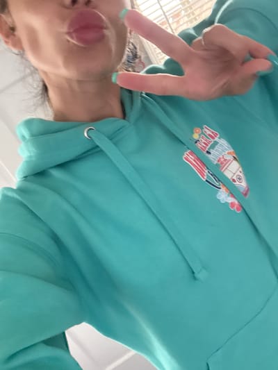 A casual, close-up image features a person wearing a vibrant teal or aqua-colored hooded sweatshirt. The person's head is visible in the lower left portion of the frame, with their tongue playfully extended from their mouth. Their left hand, positioned prominently in the upper left, displays a peace sign (V-sign) towards the viewer. A simple ring adorns their ring finger, and their fingernails are painted with a light blue-green polish.
The hooded sweatshirt features a colorful graphic design on its chest. The design appears to be a stylized, whimsical depiction of beach-related elements, possibly including a surf van or bus, and another abstract shape resembling a surfboard or flag. The graphic utilizes bright colors such as reds, whites, blues, yellows, and oranges. The hoodie also shows its matching teal drawstrings with shiny silver eyelets.
The background is largely out of focus and appears to be a light, indistinct color, suggesting an indoor setting. There are faint hints of a grid-like structure, possibly a window or vent, in the blurred background behind the hand. The overall impression is one of playfulness and casualness, captured in a candid moment. The unique angle of the shot, which is rotated 90 degrees clockwise, contributes to its informal and personal feel. No discernible text is clearly readable within the graphic or elsewhere in the image.