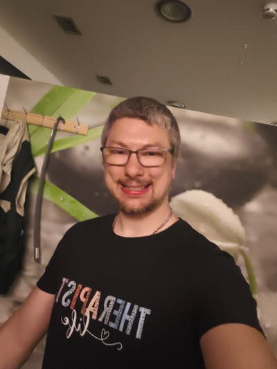This is a media file featuring a portrait of a smiling man with short, light brown hair and glasses. He is wearing a black t-shirt with colorful text that appears to say "THERAPIST LIFE" in a stylized font, with a small heart symbol below it. The man is looking directly at the camera and has a cheerful expression.

The background shows a wall with a green and white striped pattern, and a large, abstract image of a white flower or petal. To the left of the man, there are some clothes hanging on a wooden rack. The lighting suggests an indoor setting, possibly a home or a casual establishment. There are no obvious cues about the time of day or weather. The emotional tone is positive and friendly, conveyed by the man's smile. The location context provided is Troisdorf, Germany.