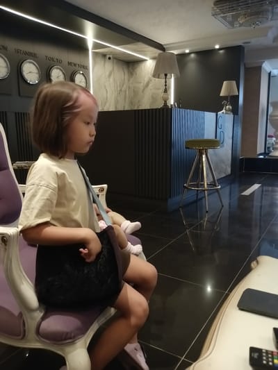 A young girl with light brown hair, dyed pink at the tips, sits on a plush purple chair in what appears to be a hotel lobby. She is wearing a light-colored t-shirt and a dark skirt, with a blue strap bag slung over her shoulder. Her legs are crossed, and she is looking to her right, with a doll resting on her lap. The lobby features a dark, glossy floor, a reception desk with a striped facade, and clocks displaying different time zones on the wall, including Istanbul, Tokyo, and New York. Two lamps with cream-colored shades sit on the reception desk. A green bar stool with a metallic base is positioned near the desk. The overall aesthetic is modern and sophisticated. It is not possible to determine the specific location within the city of Kırıkkale, Turkey, based solely on the visual information provided.