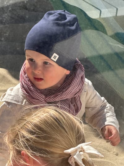 An overhead, high-angle perspective captures two young children outdoors, likely on a sunny day in Middelkerke, Belgium. The central figure, a toddler, occupies the left side of the frame, looking upwards with an engaged, slightly smiling expression and wide, light-colored eyes. This child wears a dark blue beanie featuring a white square patch with a black letter "R", a red and white striped scarf, and a light-colored long-sleeved top. Their right hand is extended in the upper right portion of the image.
To the right, only the top of another child's blonde head is visible, adorned with a white, possibly floral-shaped, hair clip. Both children are situated on a sandy, light-brown ground, consistent with a beach environment. The upper left background shows blurred, abstract patterns in shades of green and grey, which could be reflections or structural elements. The bright, natural light suggests a clear daytime setting, capturing a moment of curiosity or play.
