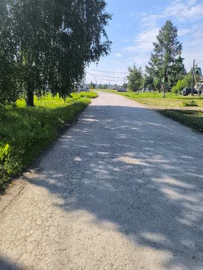 The image shows a paved road in the village of Kashino, Russia, on a sunny day. A large tree with green leaves dominates the left side of the foreground. The road is made of asphalt and shows signs of wear with visible cracks. Shadows from foliage are cast across the road. In the background, there is green grass, more trees, and a building with a green roof and gold domes, which appears to be a church. There are utility poles with wires and a few parked vehicles visible. The sky is blue with scattered white clouds.
