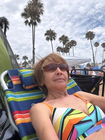 A medium shot captures a woman in a colorful bikini resting in a beach chair. She wears sunglasses and her short, brown hair is windswept. The beach chair, adorned with bright stripes of blue, green, orange, and yellow, is positioned on the sand. In the background, palm trees sway under a cloudy sky. A white canopy tent and American flags are visible further down the beach, where other beachgoers are present. The scene is set at the beach in San Clemente, California.