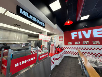 The photo shows the interior of a Five Guys restaurant, likely located in Bloomingdale, United States of America. The restaurant is in the middle of service; one employee is visible behind a stainless steel counter marked "MILKSHAKES." The order counter is lined with red and white checkered tiles, and stacks of cups and ordering equipment are present.
On the wall behind the counter, the word "FIVE" is displayed in large white letters. Numerous documents and notices are posted on the wall. Bags of potatoes are stacked in the seating area, which is marked with a wooden railing, next to the wall.
Overhead, a black ceiling features recessed lighting and a red industrial-style light fixture. Signs are visible above the counter indicating the order placement location.
