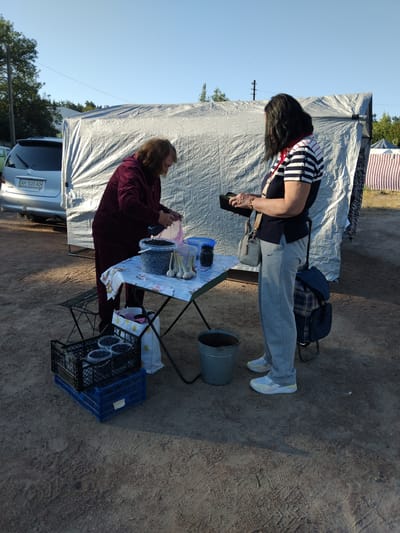 In Irshansk, Ukraine, a woman in a maroon tracksuit stands behind a table covered with a patterned cloth, pouring berries from a bag into a container. A second woman, wearing a striped shirt and gray sweatpants, stands opposite her, holding a dark object and a purse. On the table are containers of blueberries, garlic, and other items. In front of the table, to the left, are two crates of blueberries. A metal bucket is on the ground to the right of the table. A silver car is parked to the left of the scene, and a large, tarp-covered structure stands behind the people. The ground is dirt and gravel, and the sky is clear blue.