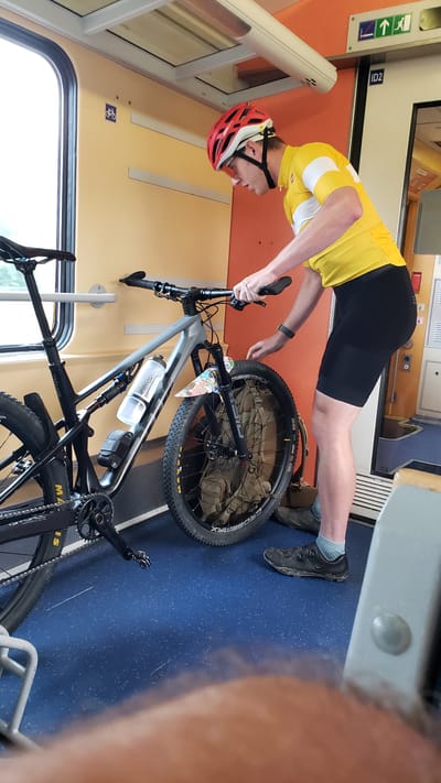 The image depicts a man in cycling attire, including a yellow and white jersey, black cycling shorts, a red helmet, and cycling shoes, standing next to his mountain bike inside a train car. The bike is gray and black, with a water bottle and a small bag attached to the frame. The front wheel of the bike has a colorful fender and a khaki-colored bag secured within its spokes. The man is looking down at the bike, with his hands near the handlebars. The interior of the train car features blue flooring and beige and orange walls. A window is visible on the left side of the image. A sign with a bicycle symbol is affixed to the wall on the left. The man's face is partially obscured by his helmet and the angle of the shot. The context suggests he is preparing the bike for transport or has just unloaded it. The image does not provide any specific visual cues to confirm the location as Liptovský Mikuláš, Slovakia.