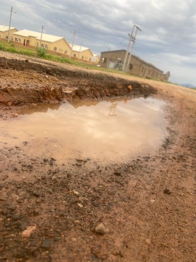 The image depicts a close-up view of a muddy puddle on a dirt road. The puddle is a light brown color and reflects the sky. The surrounding ground is composed of dirt, gravel, and small stones. In the background, there are several buildings with light-colored roofs and walls, possibly residential homes. Utility poles are visible in front of the buildings. The sky is overcast with clouds. The scene suggests it may have recently rained, creating the puddle. The time of day appears to be daytime, given the ambient light. The location is identified as Bulamare, Nigeria. No people are visible in the image.