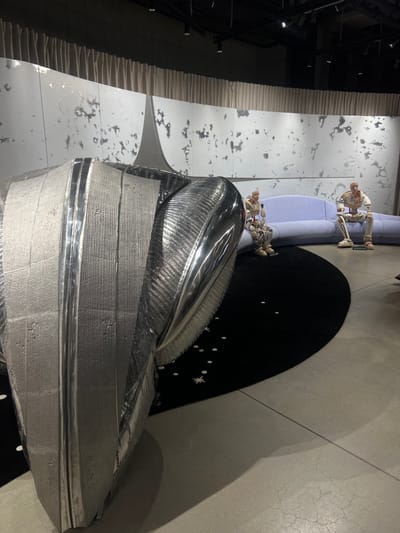 The image depicts an indoor art exhibition or installation located in 성동구, South Korea. The precise address or venue within 성동구 is not discernible from the visual information.
In the left foreground, a large, metallic silver-toned sculpture dominates the frame. It features a complex, layered form with both highly polished, reflective surfaces and textured, matte sections. The sculpture exhibits a curved, aerodynamic aesthetic.
In the right midground, two bald, humanoid mannequins are seated on a light blue, segmented, curved sofa. Each mannequin is dressed in a light-colored, multi-piece suit with metallic accents, resembling futuristic or astronaut attire. They are static, facing forward.
A large, black, semi-circular rug lies on the light gray concrete floor, positioned in front of the sofa and partially under the sculpture. The rug is decorated with scattered white circular dots and star patterns.
The background consists of a curved wall section behind the sofa, rendered in a light gray with irregular darker splotches, evoking a lunar landscape. A prominent, upward-pointing, fin-like structure is integrated into the center of this wall. Above the wall, light beige draped curtains are visible. The ceiling is dark, with exposed track lighting fixtures illuminating the scene and casting shadows on the floor. No active individuals or interactions are present.