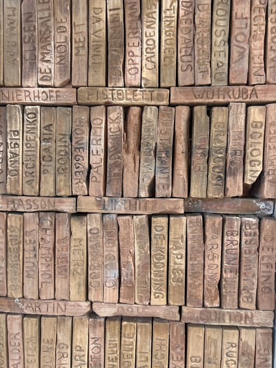 The image displays a close-up, eye-level, medium shot of a wall composed of numerous inscribed bricks. These bricks, arranged in vertical columns and horizontal rows, bear carved names and phrases, primarily in Roman capital letters. The texture of the aged bricks, with visible imperfections and variations in color, suggests they are an outdoor architectural element. The lighting is natural and slightly diffused, creating soft shadows.

The names inscribed on the bricks include recognizable figures and places such as "MAN RAY", "ELSEN", "DE MARSALLE", "NOLDE", "MEIT", "DA VINCI", "SEVERINI", "OPPERMAN", "CARDENAS", "TINGUELY", "DAEDALUS", "DARGOUT", "ROUSSEAU", "WOLF", "STEIN", "CORSE", "NIERHOFF", "PISTOLETO", and "WOIRUBA". Other inscriptions appear to be locations or names that are less universally recognized, such as "PISANO", "ARCHIPENKO", "PICARIA", "RODIN", "HONEGGER", "PALERMO", "LEADWICK", "MASSOM", "BOURGEOIS", "MULLER", "CHAMPOL", "MEIER", "MERZ", "CORNARO", "CODION", "DEBAIN", "MARTIN", "DE KOONING", "TAPIES", "BRANCUSI", "BROGNI", "ARMED", "DELEFINO", "ALDER", "IZEIZA", "DOORE", "GRIS", "ARP", "LEON", "VELLI", "DELLE", "HI", "NEN", "BI", "BURTON", and "RUC". The bricks are stacked tightly, with some slight gaps and misalignments. This arrangement suggests a decorative or memorial wall.

While the exact location is not explicitly stated in the image itself, such inscribed brick walls are a characteristic feature found in public spaces, particularly in European towns and cities as part of street art projects or historical commemorations. Given the style of names and the overall presentation, this type of installation could potentially be found in a town square, park, or along a promenade. Pollença, Spain, known for its artistic heritage and charming streets, might feature such an artistic display. However, without specific contextual information directly from the image, this remains a speculative observation based on similar installations. The forensic description is based solely on the visual evidence presented.