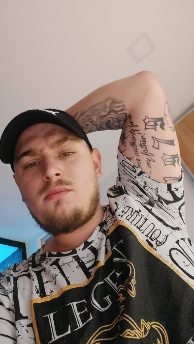 Senec, Slovakia. A close-up, low-angle portrait features a light-skinned adult male looking directly forward. He has a trimmed brown beard and mustache and wears a black baseball cap with a white emblem, and a short-sleeved t-shirt with a white, black, and gold pattern including the words "LEGEND" and "COUTURE." His right arm is bent and raised, displaying multiple tattoos: a detailed design on the upper arm, script text on the inner bicep reading "I have no authority for my father!", and stylized gothic lettering further down. The background is a plain light-colored wall with a faint square ceiling light fixture on the upper right and a blurry blue light source visible on the lower left.