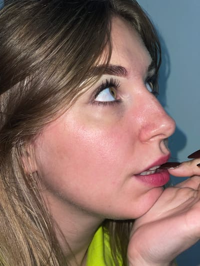 A close-up, sideways shot features the face of a person, with their head tilted upwards and to the right. The individual has long, light brown hair, some of which falls across their cheek and forehead. Their eyes are open and directed upwards, with visible eyelashes and eyebrows. The person's lips are slightly parted and appear to be red, possibly from lipstick. A finger, with dark-colored fingernails, is touching the person's mouth. The background is a plain, light blue wall, with a shadow cast upon it. The lighting suggests an indoor setting, with no clear indication of the time of day or weather. There is no visible text in the image. The scene is a close-up portrait focusing on facial features and a gesture. The setting appears to be indoors against a simple backdrop.