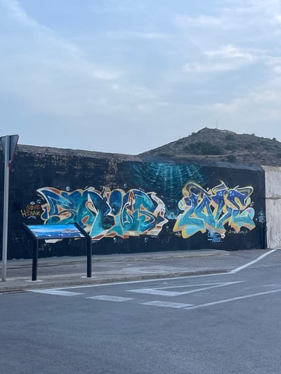 The image shows a section of a black, weathered wall featuring graffiti art in Cartagena, Spain. The graffiti is characterized by vibrant blue, yellow, and orange spray paint, with abstract lettering and bubble-like elements. One piece of graffiti reads "NOWE HINAK", while another prominent one displays stylized letters in blue and yellow. Below the graffiti, a sign with an image of a coastal landscape is mounted on two black posts. The wall is situated adjacent to a paved road with a pedestrian crossing marked by white lines. In the background, a barren, rocky hill with a few structures and antennas at its summit dominates the skyline under a hazy, overcast sky. The overall scene depicts an urban environment with an artistic element integrated into its infrastructure.