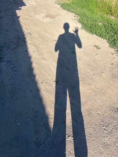 The image depicts a shadow of a person standing on a dirt path. The shadow is cast long by the sun, stretching across the ground. The person's shadow is visible from head to toe, with their left arm raised and hand open, as if waving. The path is made of dirt and small pebbles, with some tire tracks visible. To the right of the path, there is a patch of green grass. The environment appears to be outdoors, possibly in a rural or semi-rural area. The location is identified as Pereiaslav, Ukraine.