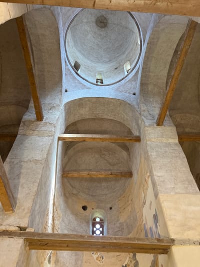 The image shows the interior of a domed structure, likely a church, with visible frescoes on the walls and wooden beams supporting the ceiling. The architecture features rounded arches and a central dome with an opening at the top. The walls are made of stone and plastered, with patches of faded frescoes depicting figures and geometric patterns. Natural light enters through a small window with a decorative grille. The scene is dusty, with cobwebs present in some areas, indicating a sense of age and disuse. The overall impression is of a historical religious building in a state of preservation. The location is identified as Spas-Nereditsy, Russia.
