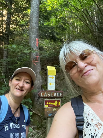 Two women are taking a selfie in a forest. The woman on the left is wearing a white baseball cap and a dark blue t-shirt. The woman on the right is wearing sunglasses and a white crocheted top. They are standing next to a wooden signpost with multiple directional signs. One sign reads "CASCADE". The background is a dense forest with tall trees and green foliage. The scene is well-lit, suggesting it is daytime. The women appear to be on a hike. There is no information about the specific location in Gumières, France.