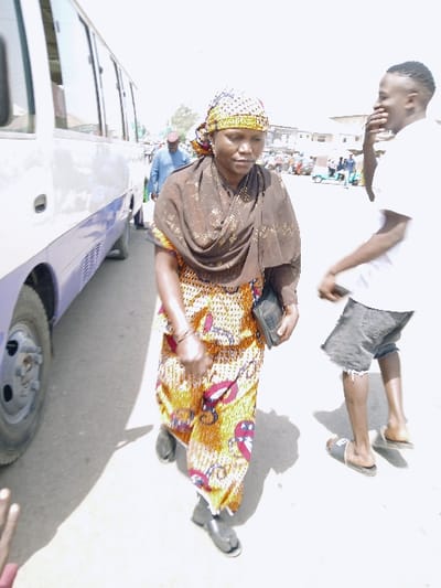 The image depicts an outdoor street scene in Mumbwa, Zambia, under bright daylight conditions. In the foreground, a woman walks towards the viewer's left. She is an adult, wearing a patterned headwrap with yellow, red, and black motifs, a dark brown patterned shawl draped over her shoulders, and a brightly colored, patterned jumpsuit or dress primarily in yellow, orange, and red hues. She carries a dark clutch bag in her left hand and wears dark, flat sandals. Her gaze is directed slightly downwards. To her right and slightly behind her, a young adult male walks towards the viewer's right. He wears a white t-shirt, dark patterned shorts, and light-colored slide sandals, with his right hand covering his mouth. In the mid-ground on the left, a white and light purple minibus or small bus is parked or slowly moving, with its front portion extending out of frame. Further in the background, another man in a blue shirt and dark red hat walks away from the camera. The ground is a light-colored surface, possibly pavement or compacted earth, with distinct shadows cast by the sun. Additional vehicles and buildings are visible in the distant background, suggesting an urban or semi-urban environment. A hand is partially visible in the lower-left corner of the frame.