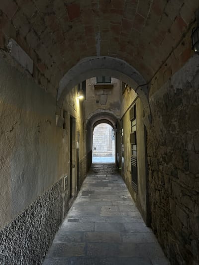The image depicts a stone-paved alleyway in Girona, Spain, characterized by an arched ceiling constructed from reddish bricks and rough-hewn stones. The alley is narrow and curves slightly, with textured walls on either side. The left wall is predominantly rendered in a pale, rough stucco, with a band of dark, pebbled material running along its base. The right wall features exposed, irregular stonework. A single bright light fixture illuminates the left wall, casting a warm glow. Further down the alley, another archway is visible, leading to an area with more stone buildings and a lighter pavement. The scene appears quiet and devoid of people, suggesting it might be early morning or late evening.