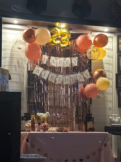 This image captures a birthday celebration setup in McAllen, United States of America. The primary subject is a decorated table positioned against a shimmering rose gold tinsel curtain backdrop. A "HAPPY BIRTHDAY" banner, with white rectangular flags featuring brown lettering and ornate borders, is horizontally strung across the tinsel curtain in two rows. Above the banner, a cluster of balloons in shades of peach, orange, yellow, and rose gold are visible, alongside clear balloons containing confetti. Centrally positioned behind these balloons are two golden foil balloons shaped to form the number "23."
The table in the foreground is draped with a light pink tablecloth adorned with small, circular rose gold polka dots. Arranged on the table are several transparent glass containers holding various snacks or candies. From left to right, these include a jar with long, thin items, another jar with red and yellow items, a martini-glass-shaped container with purple contents, and a shallow dish with light-colored items. To the far right of the table, a small dark bottle containing a dark liquid is present.
To the immediate left of the table, a tall, dark, rectangular object, possibly a speaker, partially obscures the scene. A brown Michelob Ultra beer box is partially visible behind this object. To the right of the tinsel curtain, a white slatted blind is visible, and further right, a dark, ornate furniture piece with a white dish on top is partially present. The upper wall in the background shows framed pictures. The overall lighting suggests an indoor or covered patio setting, with spotlights visible above the tinsel curtain. No individuals are present in the scene.
