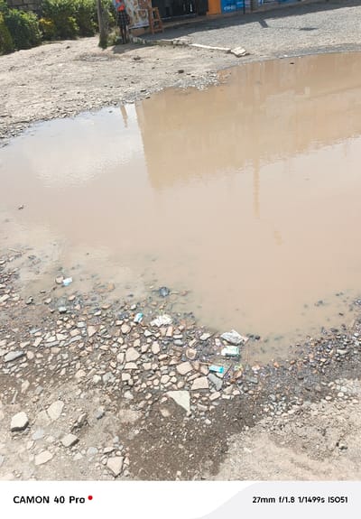 The image depicts an unpaved urban roadside in Nakuru East ward, Kenya, featuring a large, irregularly shaped body of turbid brown water that occupies the foreground and midground. The water's surface reflects a light-colored building facade and indistinct overhead elements. The perimeter of this water body is heavily littered with broken pavement fragments, small plastic containers, and miscellaneous refuse. The adjacent ground consists of compacted dirt and loose aggregate, also containing scattered waste. In the upper left background, one individual stands, wearing a dark lower garment and a lighter top. To the right, a second individual is partially visible standing near a wooden stall. Beyond the foreground, commercial buildings are present; one features a blue sign partially displaying the number '0710-533'. A dark utility pole is visible centrally in the background, and green foliage lines the far left. The scene is illuminated by daylight, highlighting the significant accumulation of standing water and refuse.