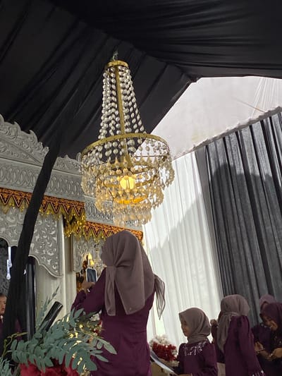 The scene depicts an interior view of an event taking place in Matangbaroh, Indonesia. A large, ornate multi-tiered chandelier, featuring a gold-toned frame adorned with numerous clear, faceted crystal or glass pendants, hangs centrally from a draped, dark fabric ceiling. The chandelier is illuminated, emitting a warm, yellowish light. The ceiling transitions to a white fabric section in the upper right quadrant of the image.

On the left side of the frame, an elaborate white wall or decorative panel is visible, characterized by intricate carvings and patterns. Below this white structure, there is a decorative border of gold and maroon fabric elements. A dark vertical column or fabric drape runs along the left edge of the white panel.

In the mid-ground, a woman is seen from behind, wearing a dark purple long-sleeved garment and a light brown or mauve hijab. She holds a white smartphone vertically in front of her, pointing it towards the decorated background, suggesting she is recording or photographing the event. To her immediate right, another woman, appearing younger, is seen in profile, also wearing a purple garment with embroidered details and a greyish hijab; she is looking towards the right side of the frame. Further to the right in the background, several other women are partially visible, similarly attired in purple garments and hijabs, some appearing to be interacting or observing. A child's face is subtly visible on the far left edge, possibly reflected or obscured.

The background on the right consists of white vertical drapes, which allow natural light to filter through, creating shadows. Beyond the white drapes, dark grey or black vertical drapes are visible. The immediate foreground on the left displays green foliage, resembling eucalyptus leaves, and a cluster of red flowers, indicative of a floral arrangement.