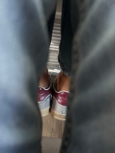 This interior image, captured in Pombal, Portugal, depicts a low-angle view between two light-colored trouser legs. In the central midground, a pair of feet wearing grey and maroon Adidas sneakers are visible from the rear. The sneakers feature light grey uppers, distinct maroon leather heel tabs with the Adidas trefoil logo, and light beige outsoles. The bare ankles of the wearer are visible above the shoes. The footwear rests on a light brown floor exhibiting a wood-grain pattern. In the background, directly behind the shoes, a light-colored surface with fine horizontal stripes is discernible, suggesting either a window blind or patterned upholstery. No specific actions or interactions are visible beyond the static placement of the feet.