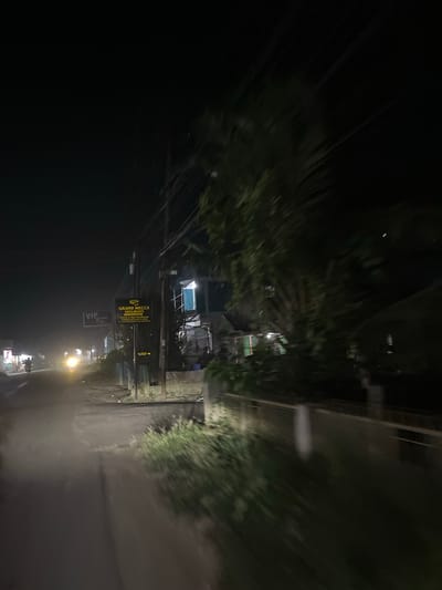 This is a nighttime, low-light, motion-blurred media file showing a street scene in Landuh, Indonesia. The perspective appears to be from a moving vehicle looking towards a street lined with trees and buildings.
The main subjects are a street, trees, buildings, and what appear to be streetlights and signs. There are hints of people and possibly motorbikes on the street in the distance, near illuminated shops. The overall scene is dark, with artificial light sources providing illumination. The motion blur suggests the image was captured while the photographer was in transit.
The time of day is clearly night. No specific weather conditions are evident, though the dust or mist on the road could suggest a dry environment or the aftermath of dry conditions. Location cues are limited to the provided context of "Landuh, Indonesia."
There are two visible signs with text. One sign, oriented vertically, is yellow with black text and reads "GRAND MECCA HOLIDAYS". Below this, another sign reads "VIP FLOX". A smaller yellow arrow points to the right, suggesting direction.
The mood is somewhat indistinct due to the low light and motion blur, but the artificial lights and roadside businesses suggest a populated area, possibly a commercial or residential street. The scene feels dynamic due to the motion blur.
