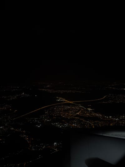 An aerial nighttime image captures the urban landscape of Rakovica Urban Municipality, Serbia. The upper portion of the frame is dark, indicative of a night sky. Below, the city is illuminated by numerous artificial lights, predominantly amber and white, forming distinct patterns across the terrain. A prominent highway system, identifiable by its bright linear illumination, features a significant curved segment and an interchange. Adjacent areas display clusters of lights consistent with urban development, interspersed with unlit regions suggesting undeveloped land or parks. A dark segment of an aircraft wing is visible in the lower right foreground, establishing the high-altitude perspective. No individuals or specific activities are discernible within the frame.