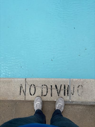 The image shows the edge of a swimming pool with clear blue water. A concrete border with the words "NO DIVING" painted in black is visible. A person wearing blue jeans and grey sneakers stands on the concrete next to the pool. The location of the image is in the city of Edison, United States of America.