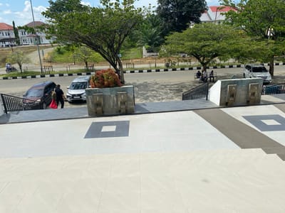 In Luengbaro, Indonesia, a man walks down a set of stairs towards two parked cars, one brown and one silver, on a paved street. He carries a red bag over his left shoulder. In the background, several buildings with red roofs and white walls are visible, surrounded by trees and greenery under a clear blue sky. A group of people sits on a bench in the shade of a large tree across the street, and a motorcycle with two riders is also seen on the road. The foreground features a modern, tiled staircase and walkway.