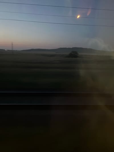 The image shows a rural landscape at dusk, viewed through a window. In the foreground, there are horizontal dark bands, possibly representing a window frame or a vehicle interior. The middle ground features a vast, flat expanse of fields, partially obscured by a layer of mist or fog that lends a diffused, hazy quality to the scene. In the distance, rolling hills rise, with the outlines softened by the twilight and atmospheric conditions. A single, small tree stands prominently on one of the hills. A tall, dark silhouette of a pylon or communication tower is visible on the far left horizon, with faint lights near its base. The sky above transitions from a pale, dusty pink near the horizon to a deep, muted blue overhead. Three thin, horizontal lines, suggestive of power lines, traverse the upper portion of the sky. A bright, yellowish-orange reflection, possibly from an interior light source, is visible on the upper right, superimposed on the sky and the power lines. The overall impression is one of quiet stillness and the muted colors of early evening in a rural setting.