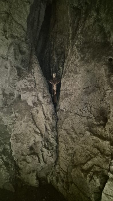 A crucifix with a figure hangs precariously within a cavernous space, wedged between rocky ledges. The cavern walls, textured with rough, uneven surfaces, dominate the scene. Light and shadow interplay across the rocks, highlighting their jagged edges. The crucifix is the focal point, contrasting against the harsh, natural environment.