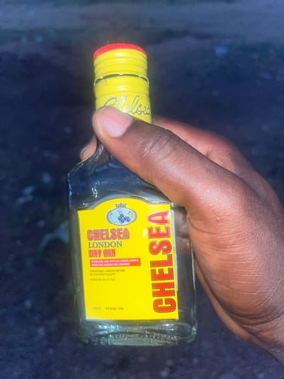 A person's dark-skinned hand is holding a small bottle of Chelsea London Dry Gin. The bottle has a yellow label with the word "CHELSEA" in red capital letters. The label also indicates that the gin is produced and bottled under license from The Chelsea Gin Company. The bottle has a yellow and red cap. The background is blurred and dark, but appears to be an outdoor setting. Based on information on the label indicating "NAFDAC REG. NO.: 01-1456," which is a registration number for the National Agency for Food and Drug Administration and Control in Nigeria, the image is likely taken in Nigeria, potentially Offatedo.
