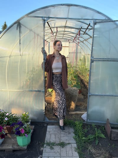 A woman stands in the open doorway of a greenhouse. She is looking to her right, out of the frame, and her right hand rests on the edge of the greenhouse frame. She is wearing a brown open shirt over a beige tube top and black and white zebra-patterned pants. She has on black shoes. Inside the greenhouse, rows of plants, possibly tomatoes, are growing. Straw bales are visible on the ground. Outside the greenhouse, to the left of the entrance, are several potted flowers. The greenhouse is constructed of a metal frame covered in corrugated plastic sheeting. The ground in front of the greenhouse is paved with light-colored rectangular stones. The background shows a clear blue sky and a hint of a tree. The overall scene appears to be a domestic garden setting. This location is in Kansk, Russia.