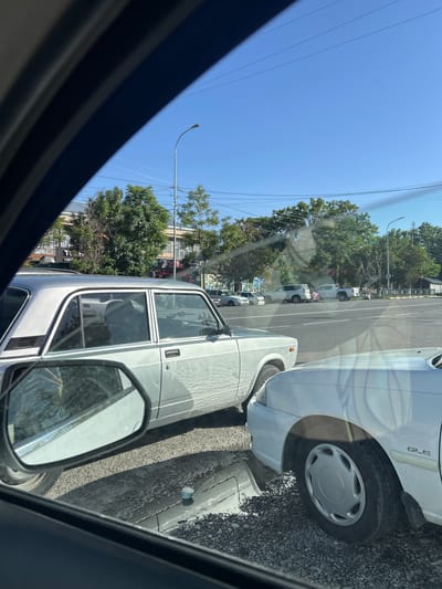 The image shows a view from inside a car, looking out onto a street in Аксукент, Kazakhstan. The scene includes several parked cars. A white car with the letters "GLE" visible is prominent. Another car is beside it. A side mirror is in view. In the distance are trees, buildings, and parked cars. The sky is blue with power lines visible. The ground appears to be asphalt and gravel. A light post is visible.