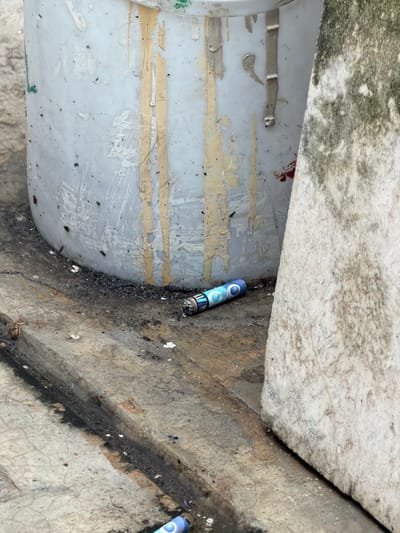 A dirty, cluttered space features two blue and white firecrackers discarded on a cracked concrete surface. Blackened debris and grime surround them. The concrete is stained with a dark, oily substance. Adjacent to the concrete, a peeling, aged white object is visible, adorned with remnants of adhesive and various marks. Moss or mold grows on an overhead surface, adding to the unkempt and neglected appearance of the scene.