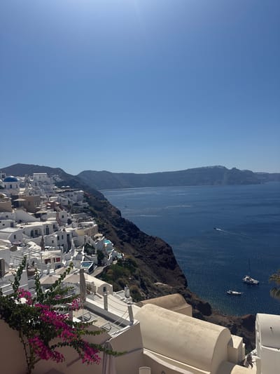 This image shows a panoramic view of the village of Oia in Santorini, Greece. The iconic white-washed buildings with blue domes cascade down the hillside towards the Aegean Sea. The sea is a deep blue and several boats are visible, including a sailboat and a smaller motorboat. The sky is clear and bright blue. In the foreground, a bougainvillea plant with vibrant pink flowers adds a splash of color to the scene. The architecture is characteristic of Cycladic style, with cubic houses, narrow pathways, and domed churches. The village is built on the caldera cliffs, offering breathtaking views of the surrounding volcanic islands. The scene is bathed in bright sunlight, suggesting a warm, sunny day. There are no people visible in the image.