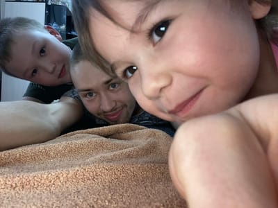 From an inverted perspective, the image shows three individuals and a textile object. A young child's face is in the foreground, occupying a large portion of the frame, partially obscured by hair. An adult male is positioned behind the child, his face visible. Another child is visible to the side of the man's face. The textile is above their heads. The location appears to be indoors, but the background is too blurry to discern specific details. There is no indication of Bely Klyuch, Russia.