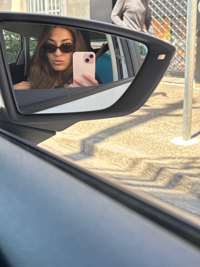 A woman with brown hair, wearing sunglasses, takes a selfie in a car's side mirror. She is holding a pink smartphone in her right hand, angled to capture her reflection. The reflection also shows part of her body, wearing a dark top, and the interior of the car, including a blue headrest. Outside the car, the reflection shows trees and a building with windows. The side mirror itself is black. The background of the original image shows a paved area with shadows, a metal fence, and a grey pole. The context of Girona, Spain, is not visible in the image.