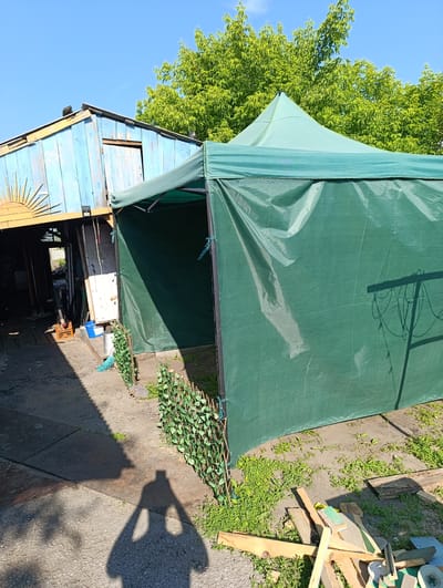 In Chernakovo, Russia, a rural scene unfolds under a clear blue sky, dominated by a large, dark green pop-up tent situated in what appears to be a yard or workshop area. The tent is set up on a concrete and gravel surface, with sparse green grass and weeds growing around its base. A small, decorative fence made of artificial greenery stands in front of the tent. To the left, a weathered, light blue wooden structure with a sloping roof is visible, featuring a rustic wooden door and an orange sunburst decoration. Various items are scattered around the area, including a blue bucket, a broom, orange plastic crates, lumber scraps, and a circular saw blade. The shadow of a person holding a camera is cast prominently on the ground in the foreground, indicating the perspective of the photographer. In the background, lush green trees provide a natural backdrop. A small, light-colored animal, possibly a dog, can be seen inside the entrance of the blue structure.